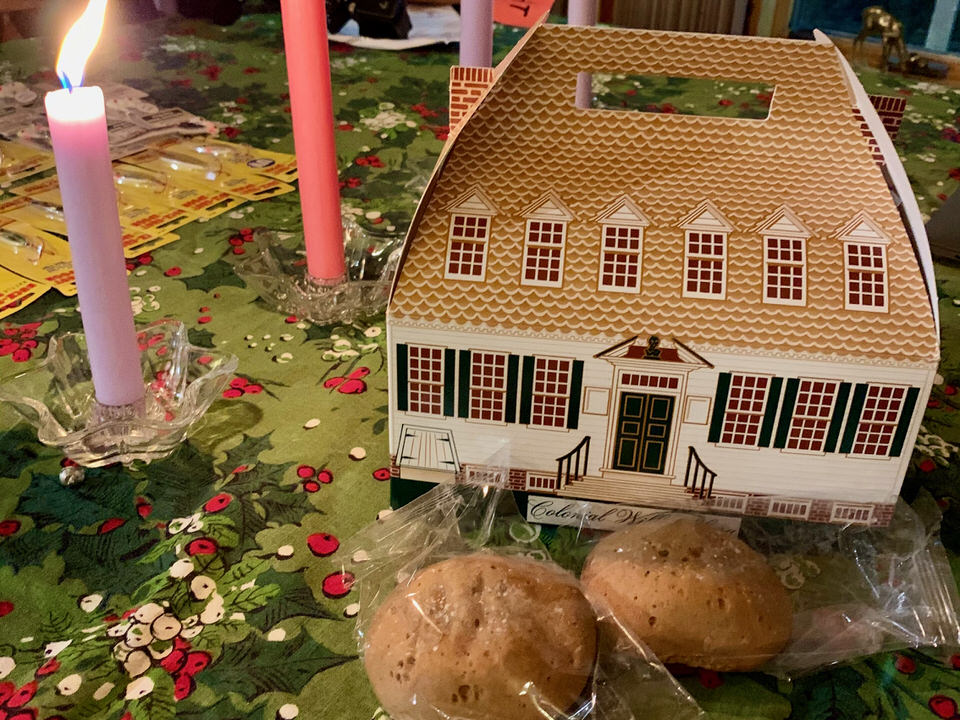 Ginger cakes and a decorative book that looks like a Colonial Williamsburg building sits on a tablecloth next to a lit taper candle