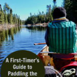 Man wearing a green lifejacket sits in a Kevlar canoe in the Boundary Waters Canoe Area Wilderness. Text overlay reads "A First-Timer's Guide to Paddling the Boundary Waters."