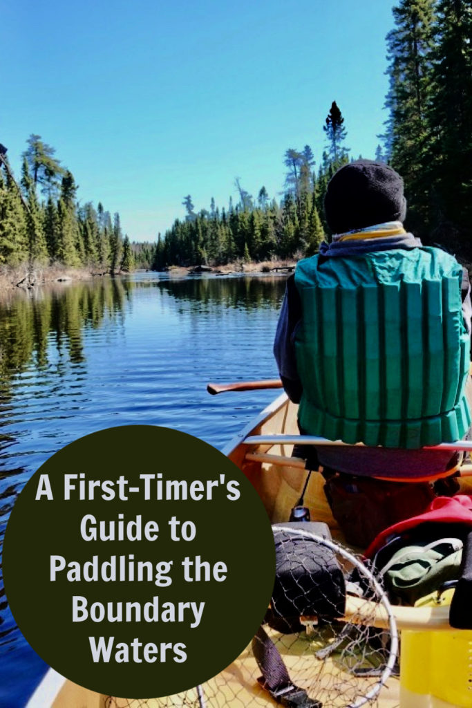 Man wearing a green lifejacket sits in a Kevlar canoe in the Boundary Waters Canoe Area Wilderness. Text overlay reads "A First-Timer's Guide to Paddling the Boundary Waters."