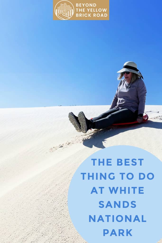 Woman slides down a white sand dune. Text overlay reads "The Best Thing To Do At White Sands National Park."
