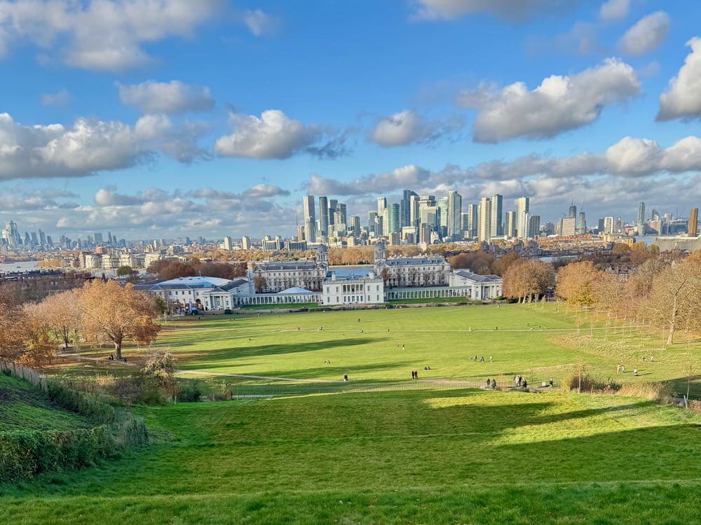 Looking across a green park expanse towards Old Royal Naval College Greenwich, the Thames River, and Canary Wharf