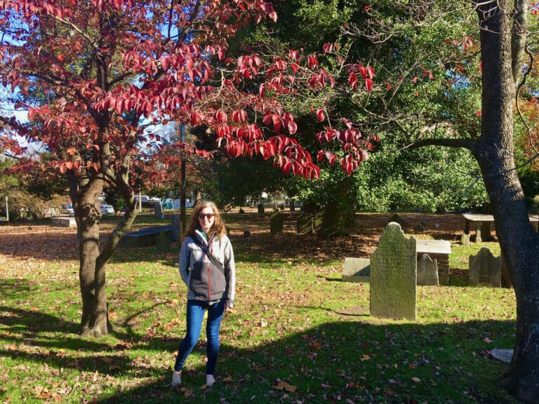 Woman standing in front of a 18th century gravestone and tree with red autumn leaves in downtown Richmond Virginia