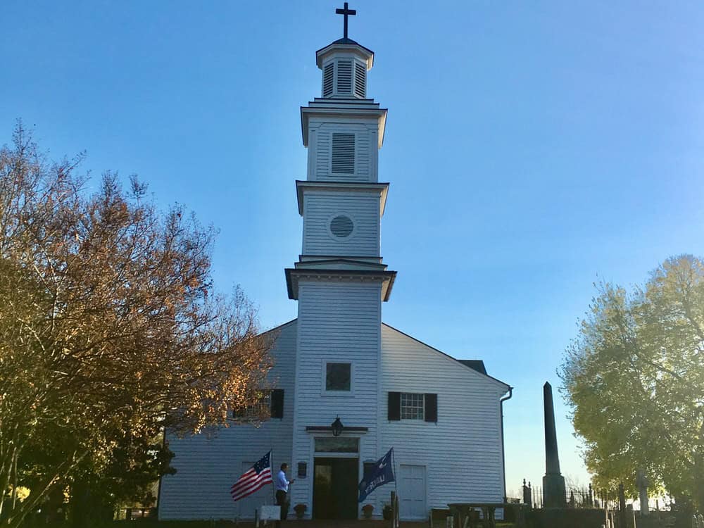 Small white church with four tiered spire framed by trees with autumn leaves. A small crowd gathers outside.