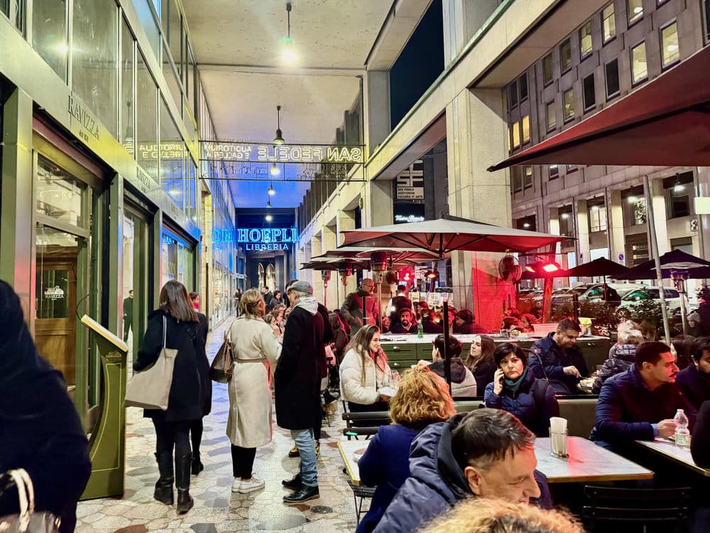 Brightly lit outdoor seating area in Milan Italy with aperitivo patrons seated at tables in front of a restaurant and pedestrians strolling past