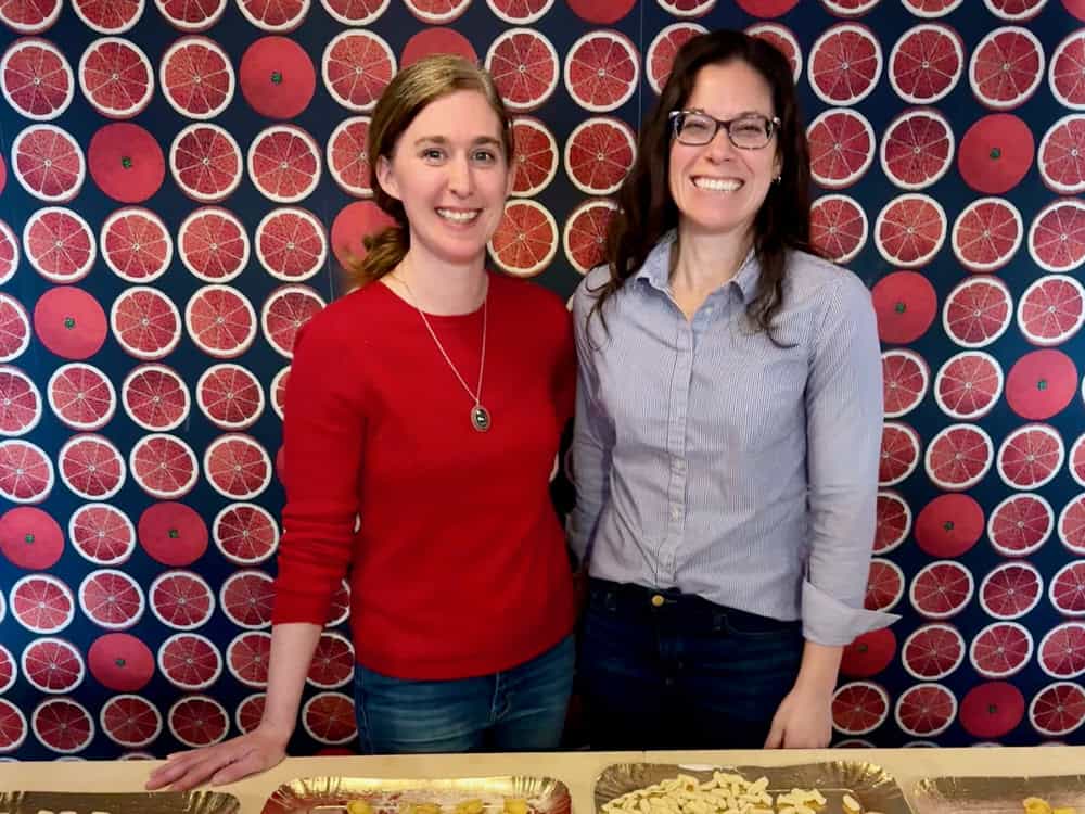 Two women standing in front of a decorative wall posing with homemade pasta