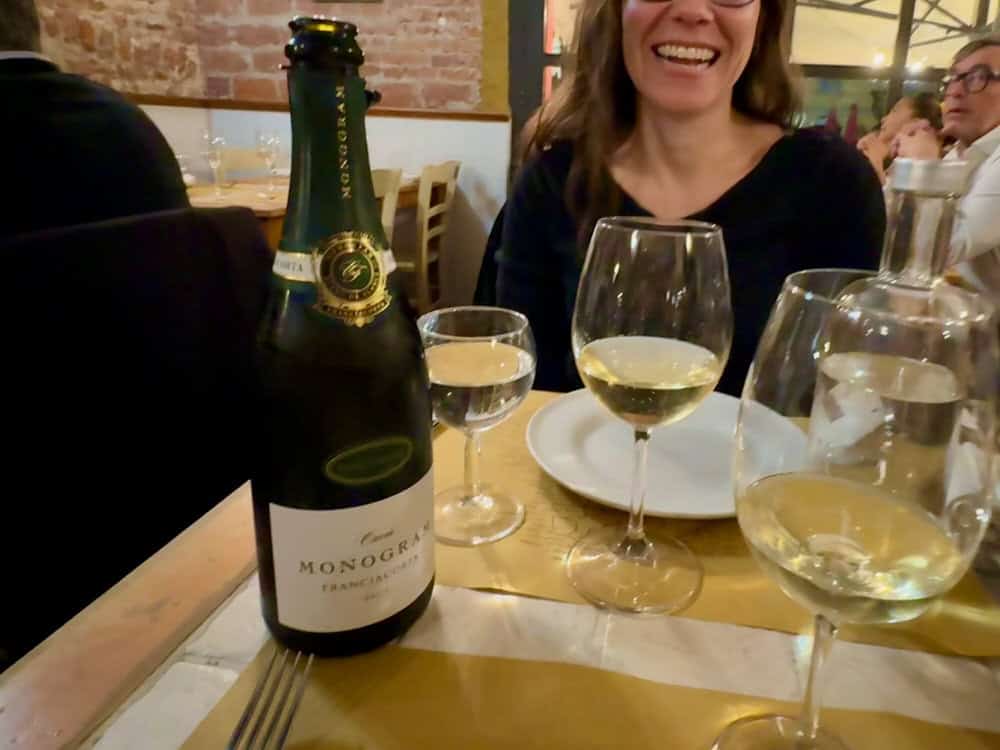 An open bottle of franciacorta sparkling wine sits on a Milan restaurant table next to table settings and glasses of wine and water. Diners are visible in the background.