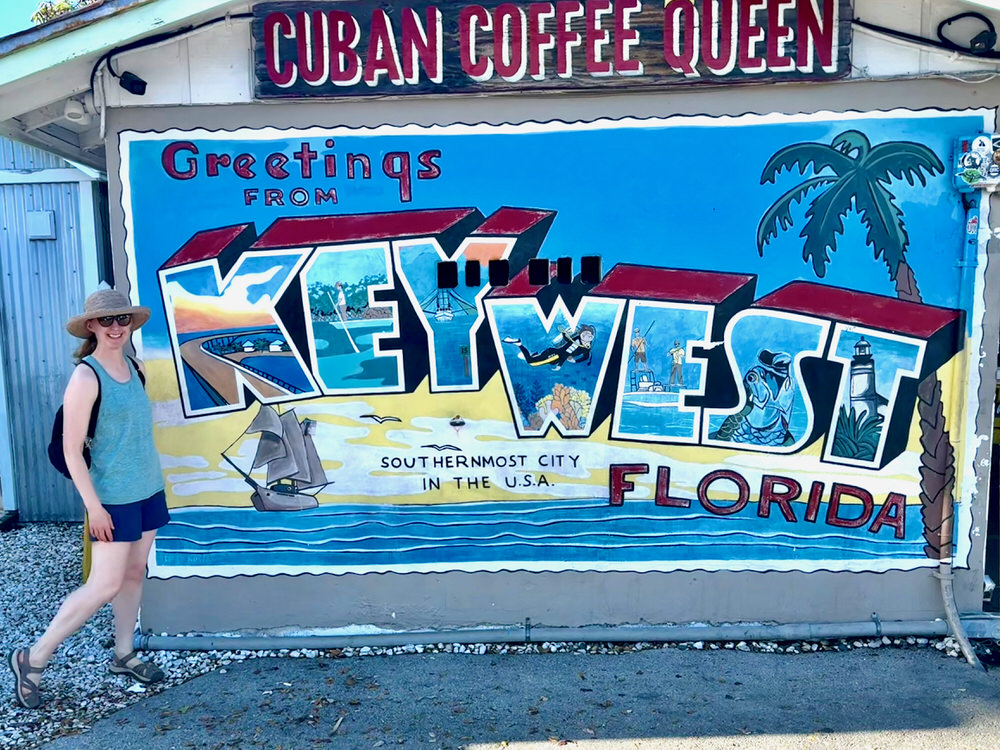 Woman wearing shorts and tank top poses in front of the postcard style mural that reads "Greetings from Key West, Florida. The Southernmost city in the USA."