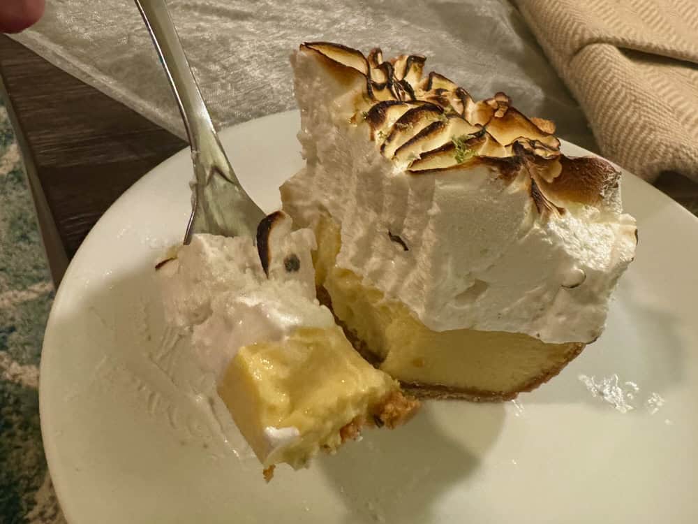 A fork holds up a bite of key lime pie. A half eaten piece of pie sits on a white plate behind the fork. Pie has pale yellow filling and a toasted meringue topping.