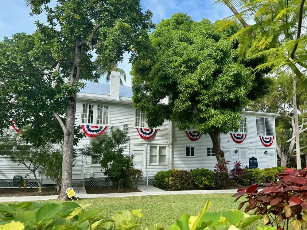 Exterior of Truman Little White House as seen through a grove of trees. Windows of the building are lined with American flag bunting.