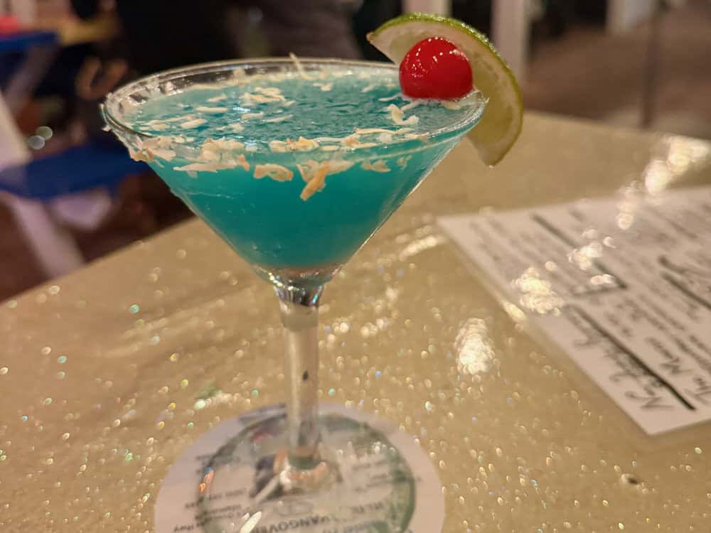 Bright blue alcohol drink in a martini glass topped with toasted coconut and a lime wedge and cherry on the rim