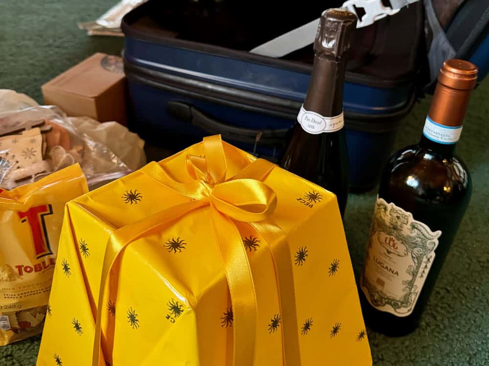 Gift packaged Milanese pandoro and Lombardy white wine in front of an open suitcase.