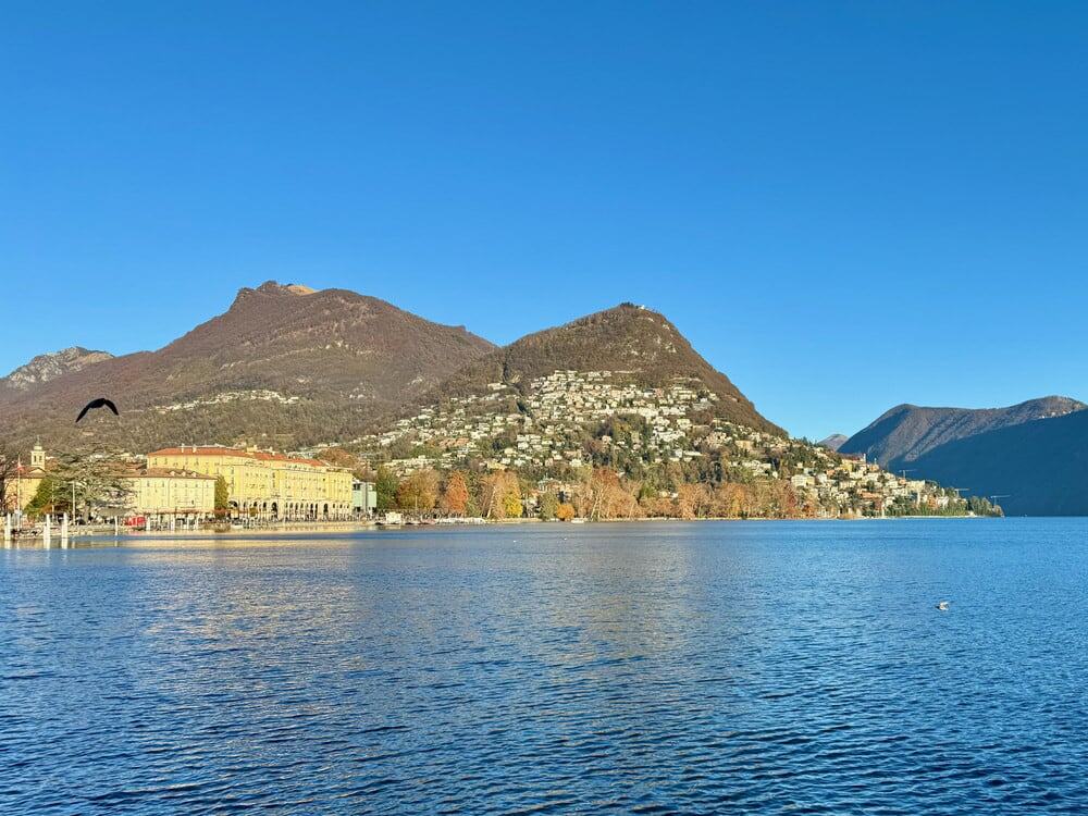 Two mountain peaks, as well as serve buildings are seen from across a large expanse of water in Lugano Switzerland