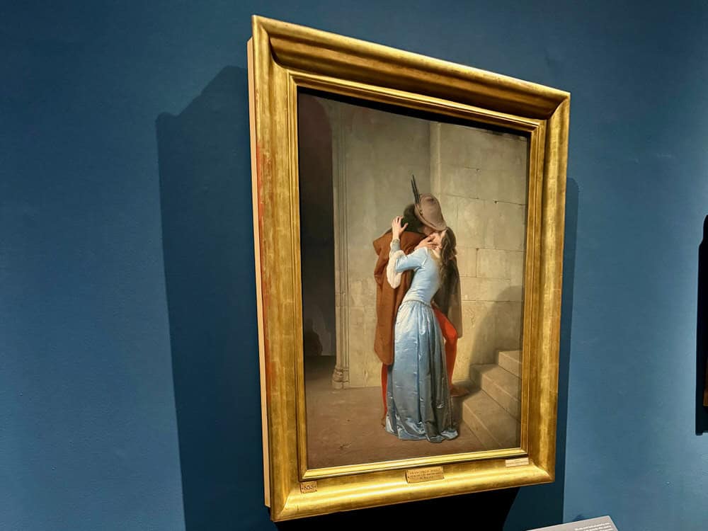 Original painting of The Kiss by Francesco Hayez hangs in a gold frame on a blue painted wall in Milan's Pinacoteca di Brera art gallery