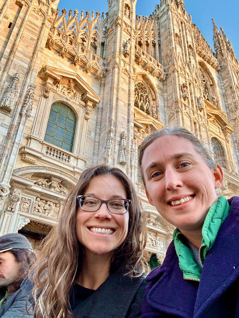 Two women take a selfie in front of the white marble facade of Milan's Duomo cathedral.