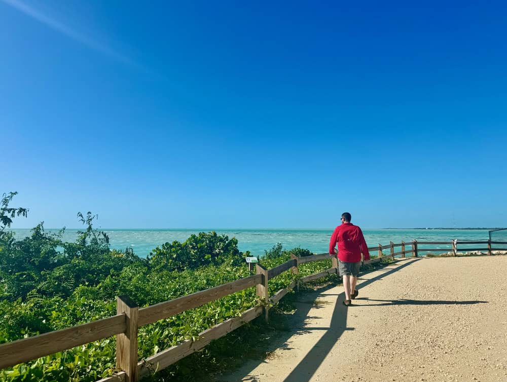 Man wearing a red shirt and shorts walks on a wide dirt pathway near green vegetation and the ocean in the Florida Keys.