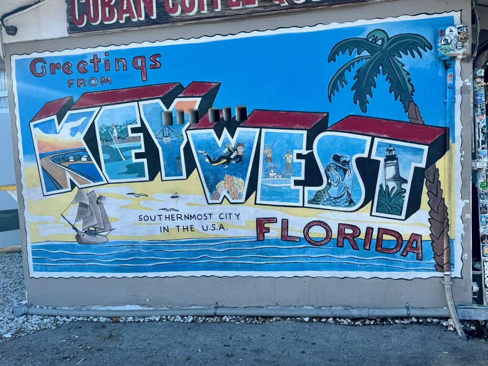 Blue mural that reads "Greetings from Key West Florida" and depicts scenes of everyday life in the Florida Keys including sailboats and palm trees.