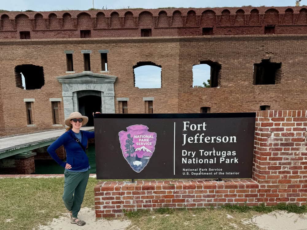Woman stands in front of the Dry Tortugas National Park Fort Jefferson sign in front of massive brick fort in the Gulf of Mexico