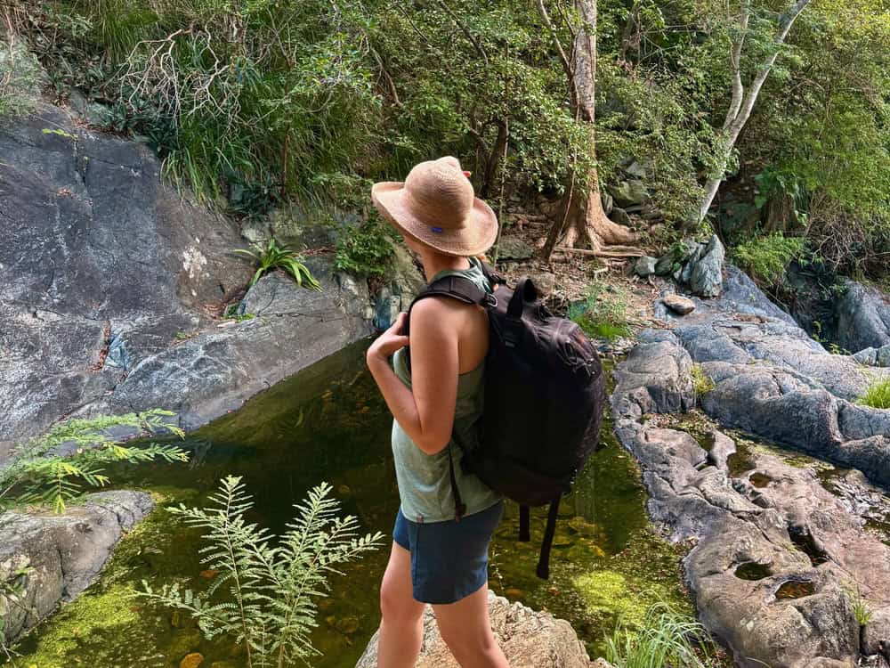 Woman wearing a backpack looks across a small pool of water on a rocky tropical trail