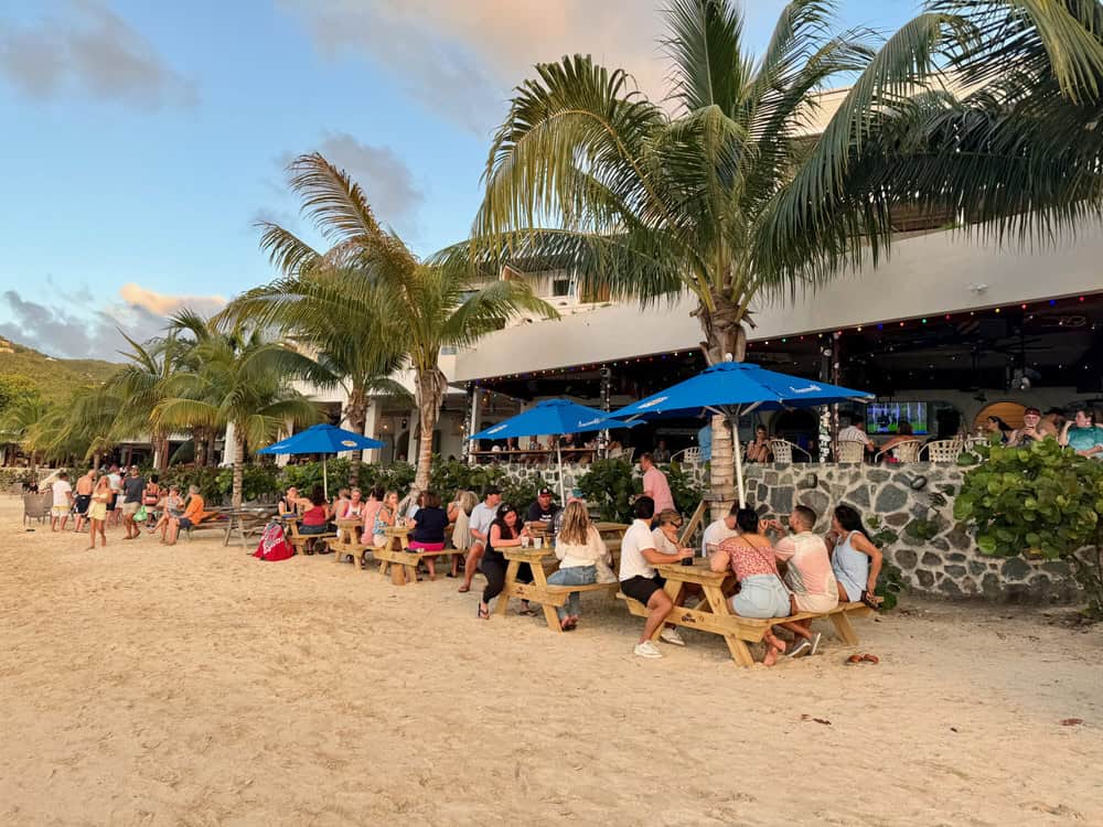 Beach bar guests sit on picnic tables on a sandy beach in Cruz Bay.
