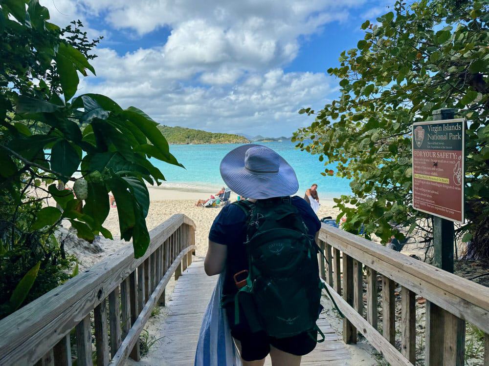 Woman wearing a backpack and sunhat walks through tropical foliage to a sandy beach