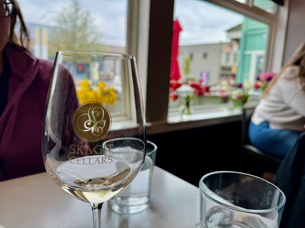 Glass of white wine on a wooden table top in front of a large window looking out on a small down town in the Pacific Northwest.