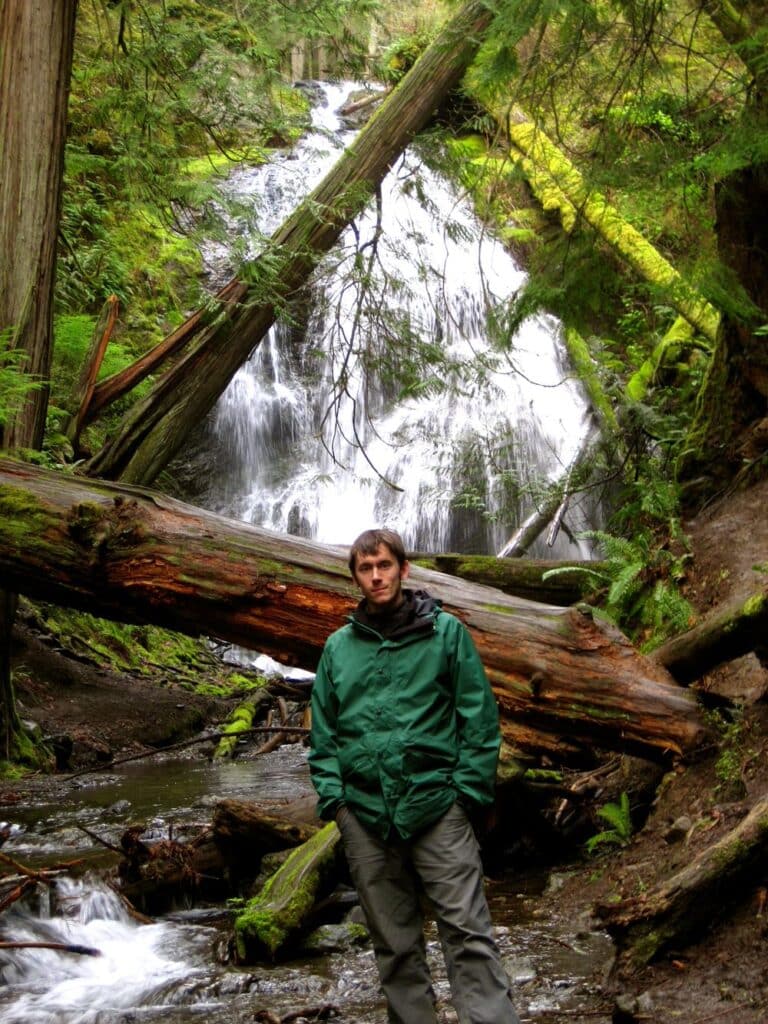 Man stands in front of a fallen tree trunk in front of a waterfall