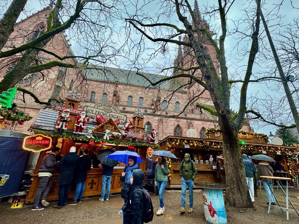 Shoppers with umbrellas stroll through a rainy Christmas market in front of a sandstone cathedral in Basel, Switzerland