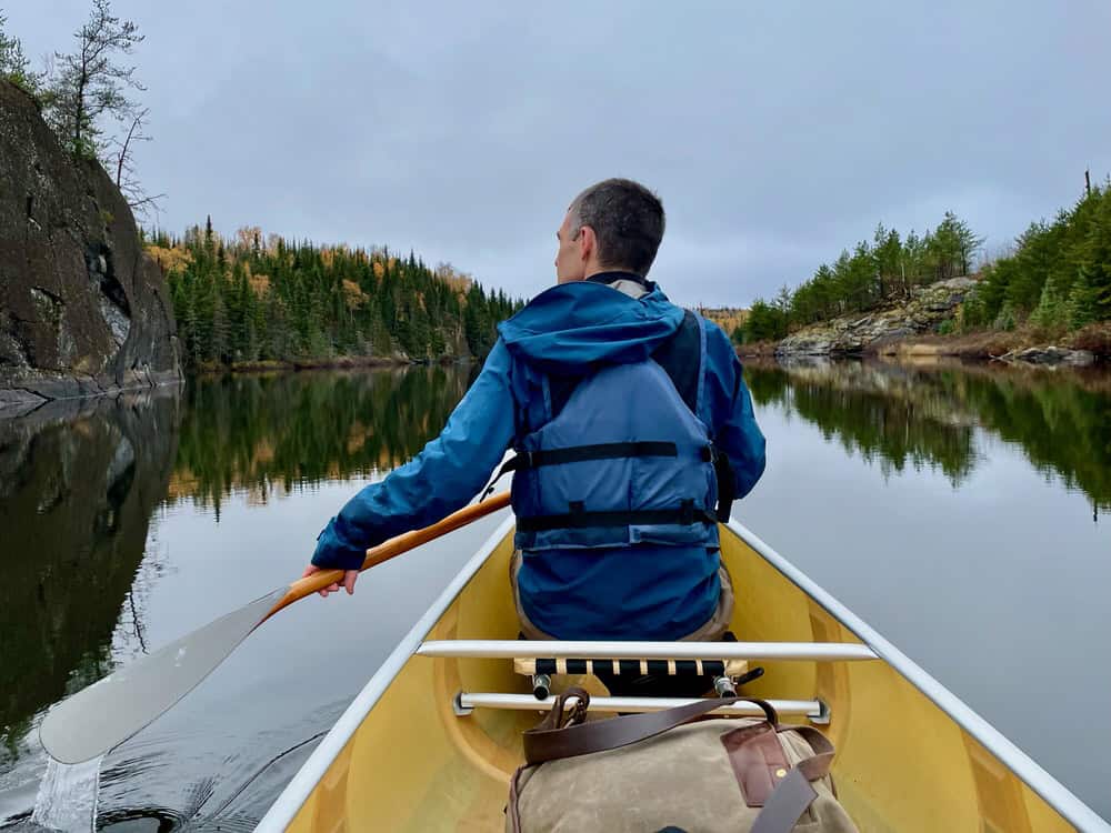Man wearing a blue rainjacket and life jacket paddles a yellow kevlar canoe on a still wilderness lake in northern Minnesota
