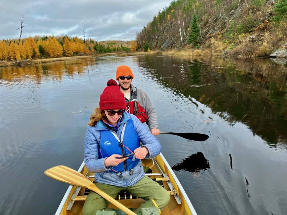 Man and woman sit in the back of a canoe on a still lake lined with fall foliage.