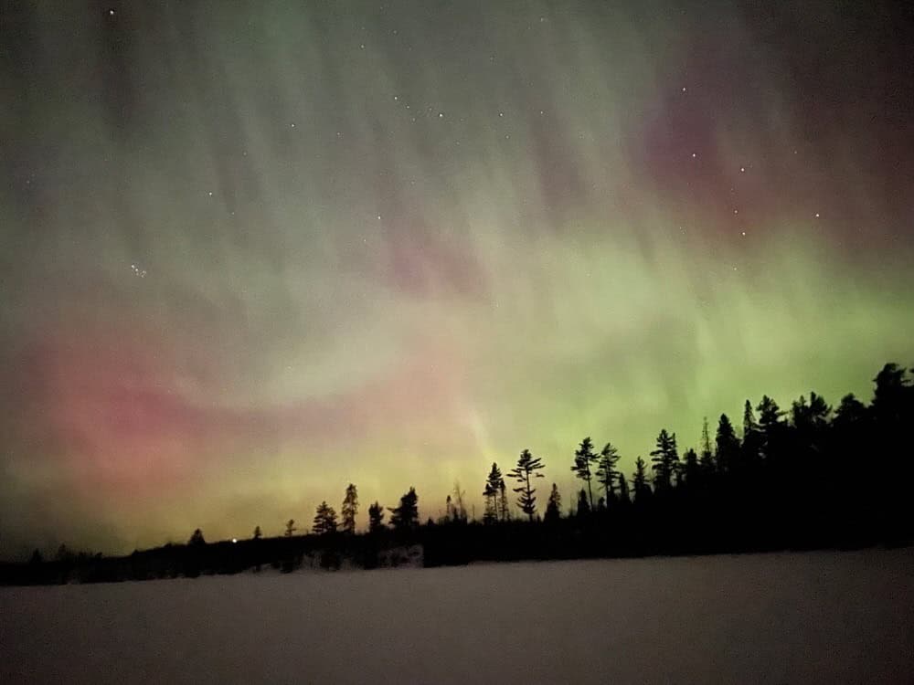 Purple and green aurora borealis over a dark northern Minnesota tree line as seen from a snow covered lake.
