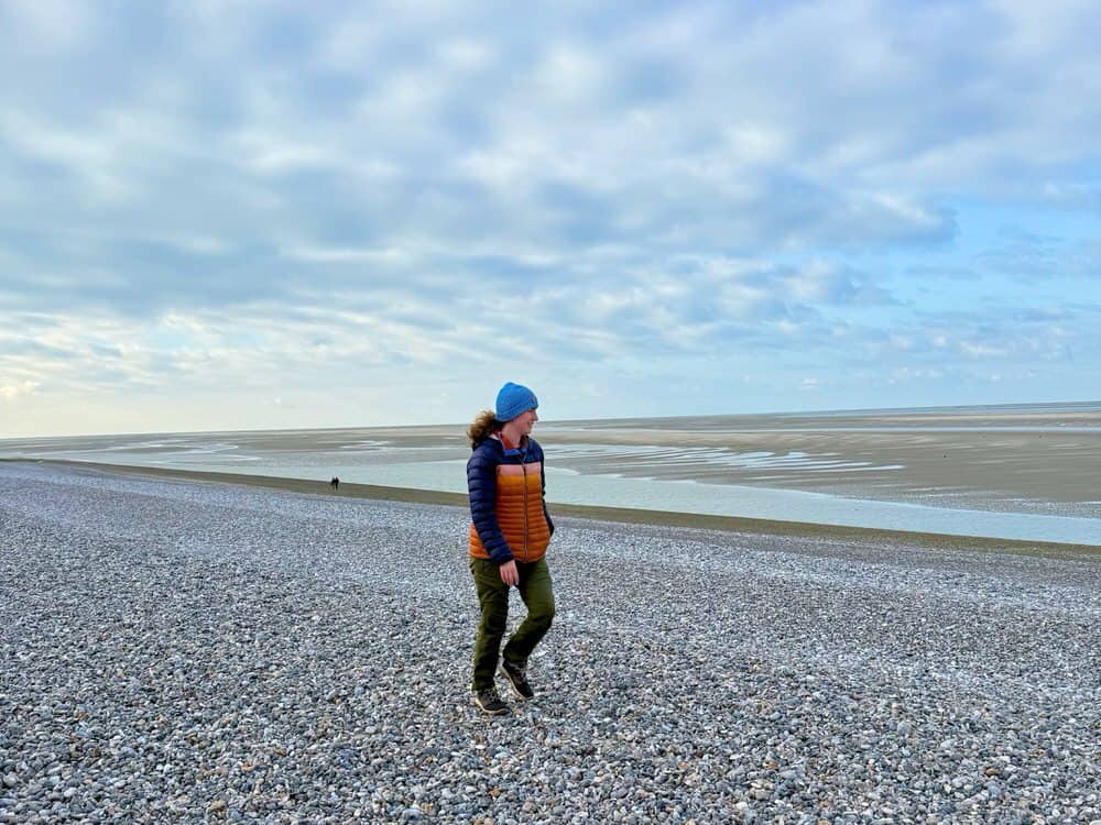 Woman walks along a large pebble beach at low tide.