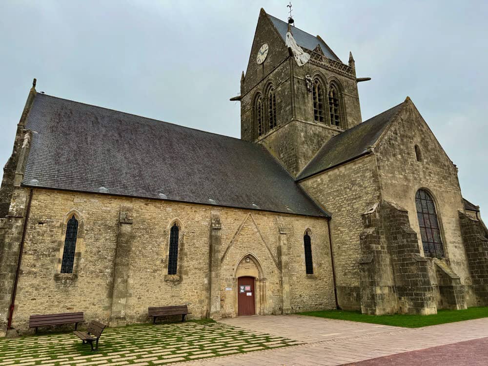 A small stone church with a mannequin paratrooper hanging from the clock tower.