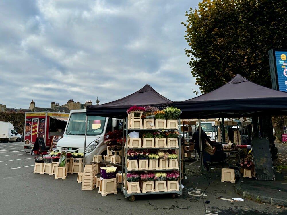 A display of flowers next to an open air market stall tent in a Bayeux parking lot