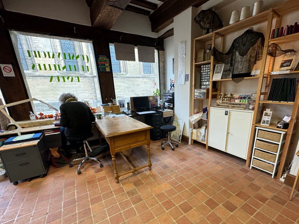 A woman sits in front of a large picture window in a lacemaking studio.