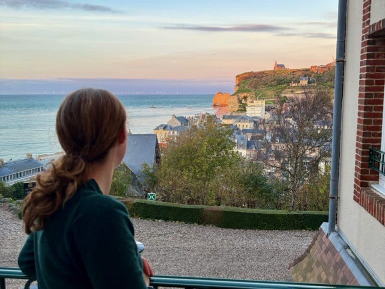Woman looks towards Etretat's upstream cliff from a hotel balcony.