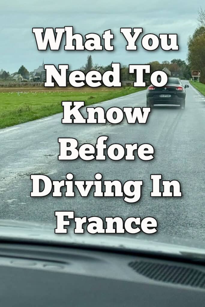 Photo taken through a car windshield looking down a country lane traveling towards Mt. St. Michel in Normandy, France. Text overlay reads "What You Need To Know Before Driving in France."