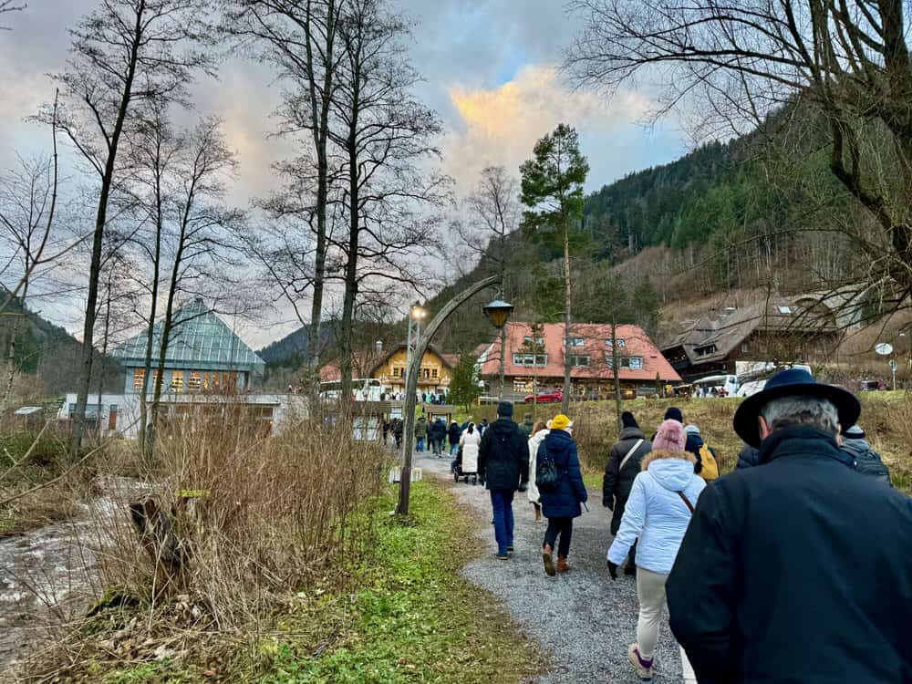 A line of people wearing winter coats hike on a gravel path to buildings on the edge of the German forest