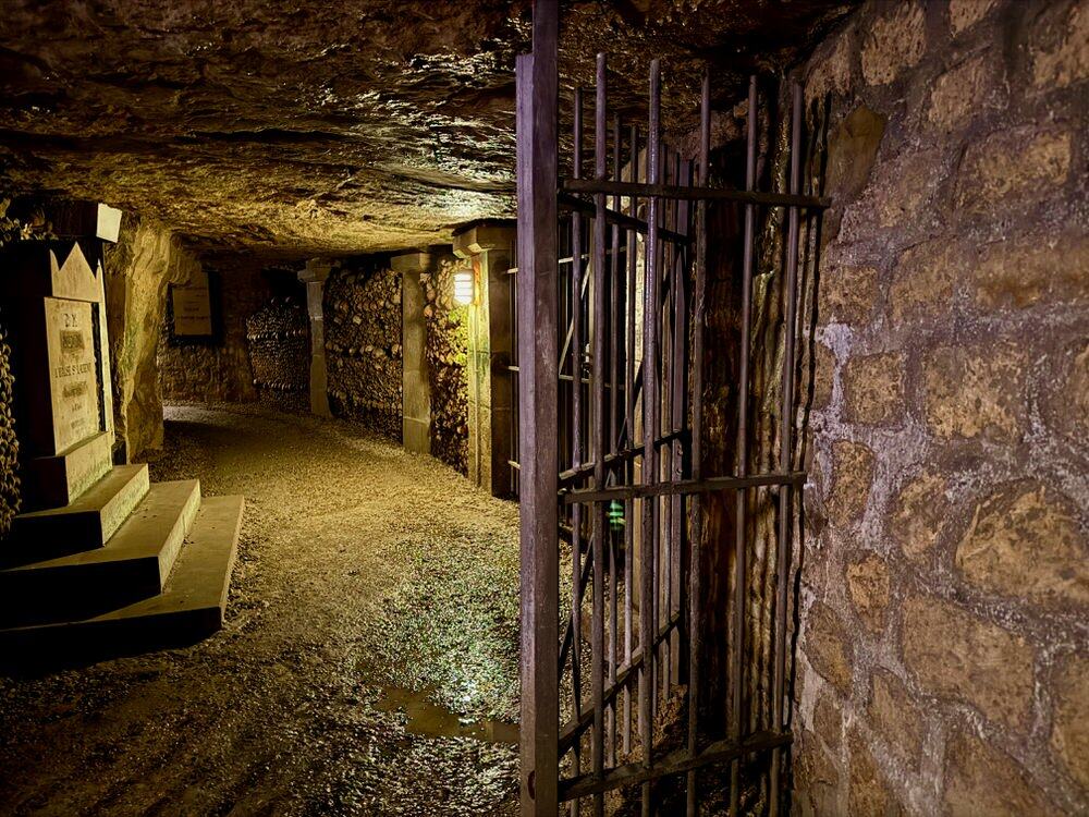 A dimly lit underground tunnel lined with human bones as seen through a metal gate.