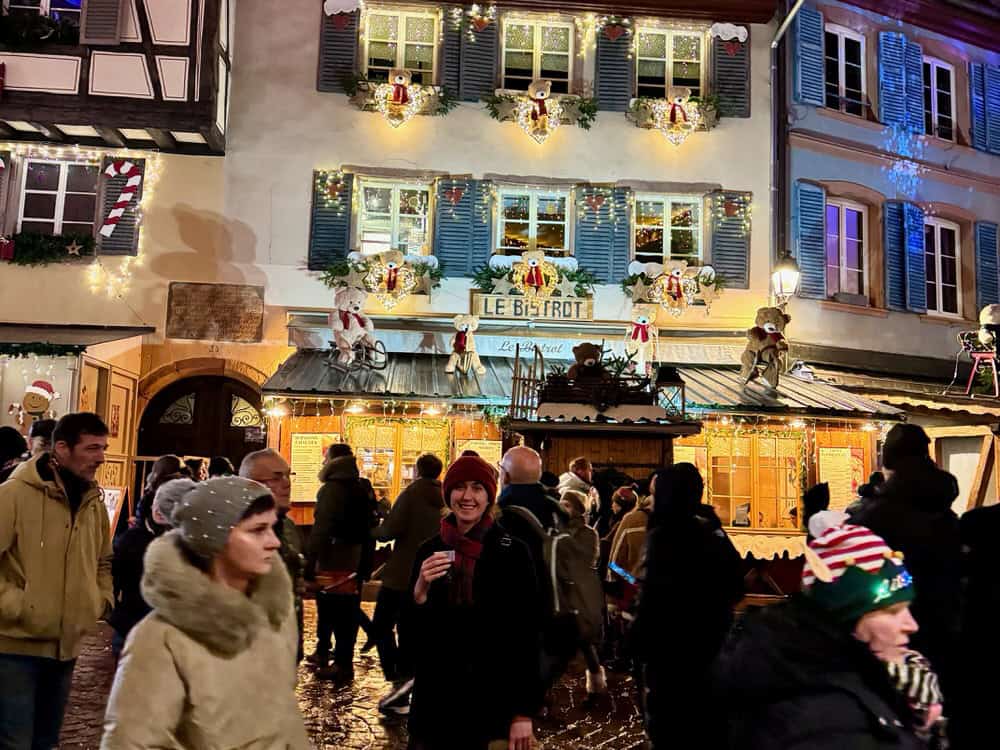 People walk around a woman holding a glass of vin chaud in front of a decorated French bistrot in Colmar
