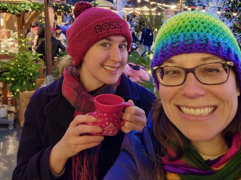 Two woman smiling in a French Christmas market holding a souvenir gluhwein mug shaped like a boot