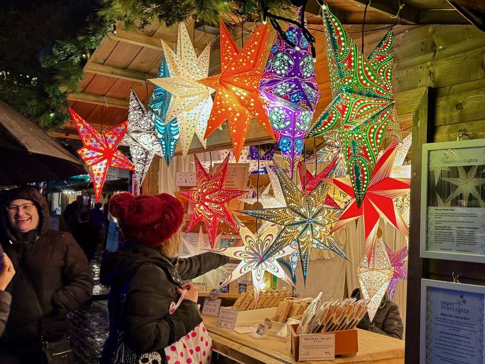 A woman stands in front of a small wooden stall strung with multicolor star shaped paper lanterns at a Christmas market in York England