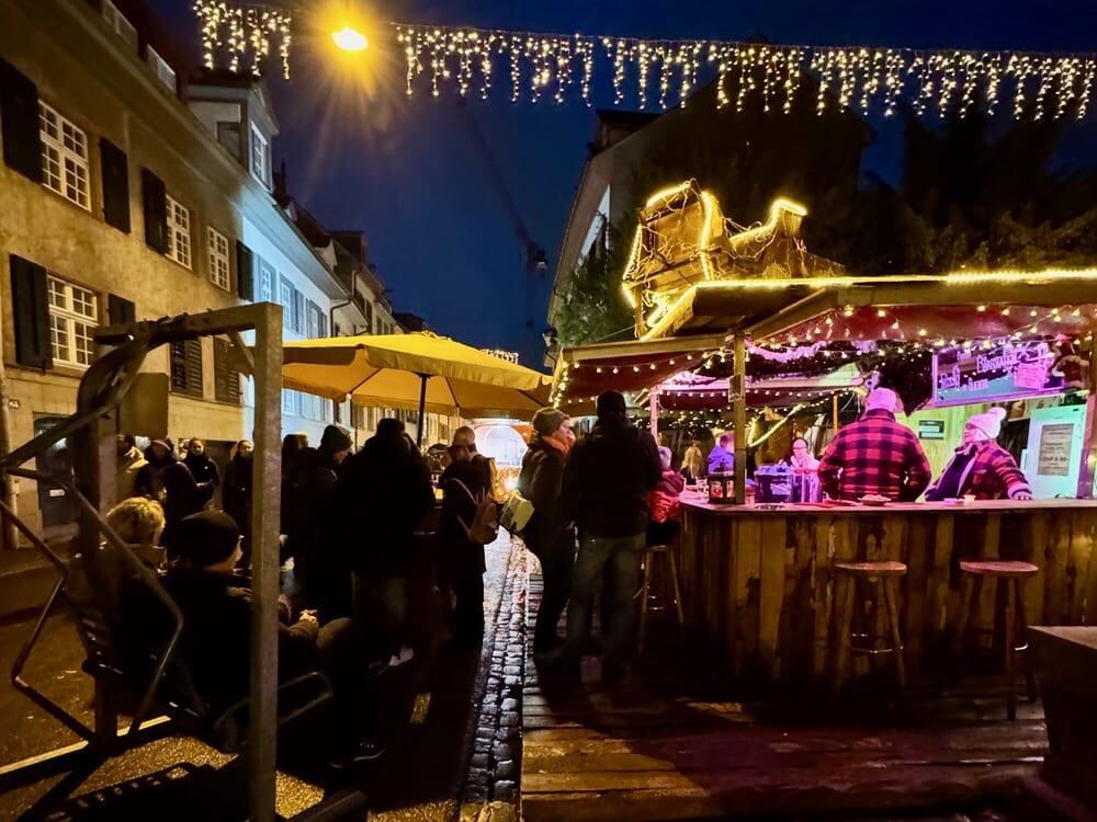 People gather outside a brightly lit bar stand after dark in Basel Switzerland's Advantgaas Christmas market