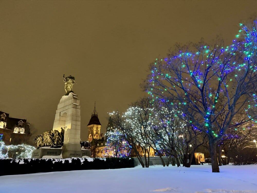 The Canada National War Monument lit at night surrounded by Christmas light covered trees in the snow.