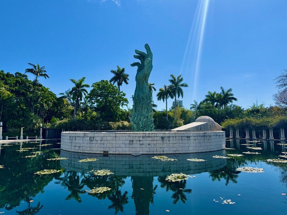 A cover hand statue with human figurines climbing up stands in the center of a pool surrounded by palm trees in Miami Beach