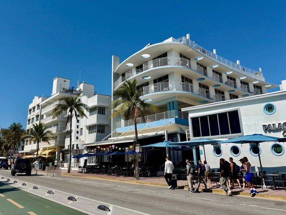 A group of people walk past the white and blue art deco exterior of Palace Bar and Restaurant on Ocean Drive in Miami Beach.