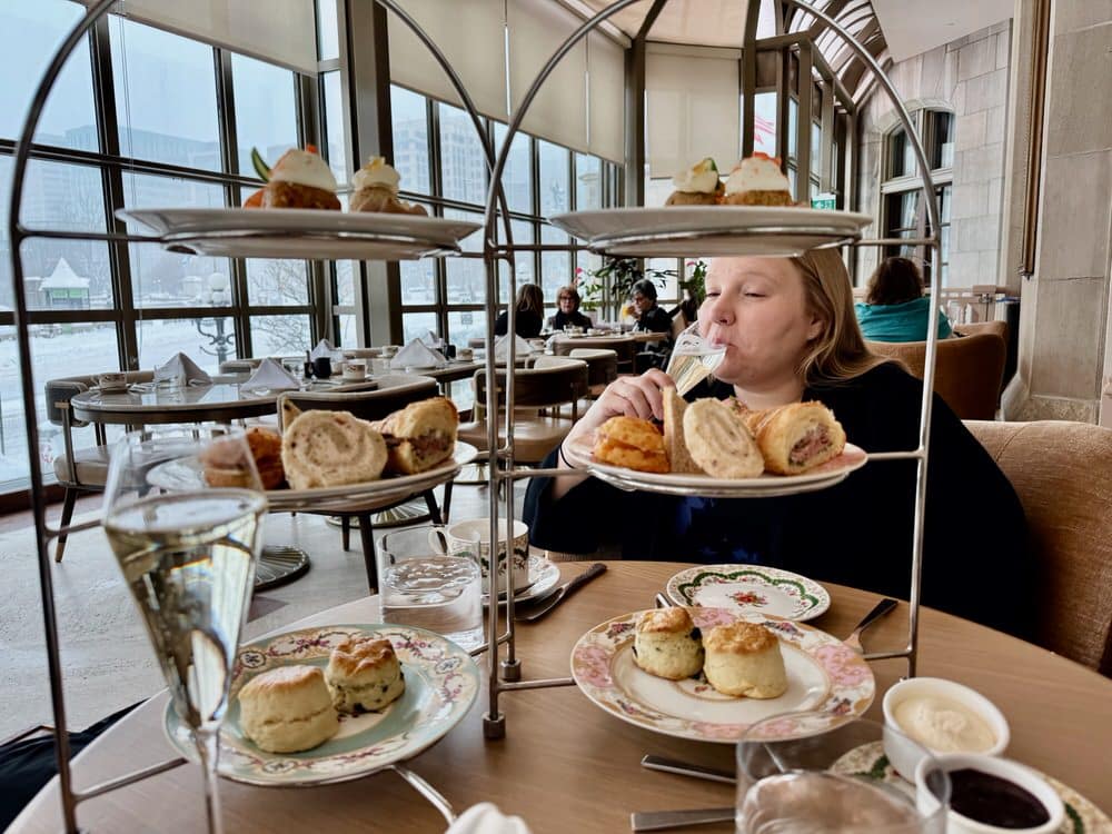 A woman sips champagne behind two trays filled with afternoon tea sandwiches and sweets