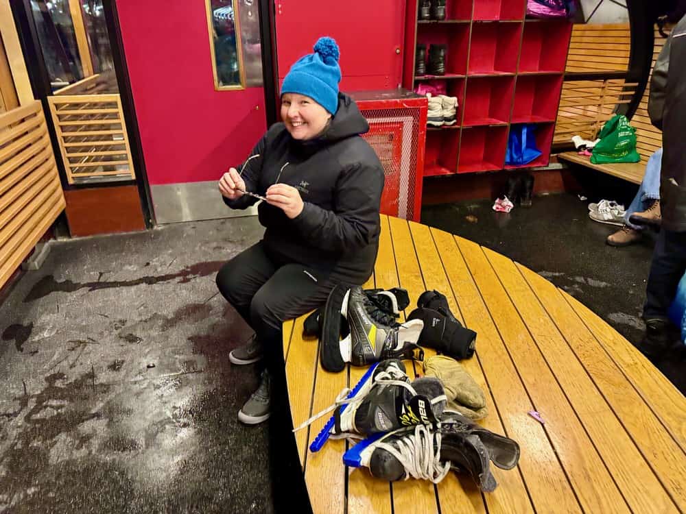 Woman sits on a bench in a warming house next to a couple pairs of hockey skates.