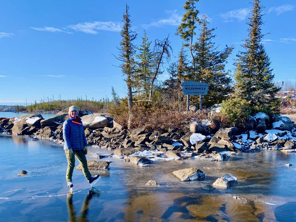 Woman wearing hockey skates poses in front of a Boundary Waters Canoe Area Wilderness sign on Seagull Lake during a wild ice skating season in early December