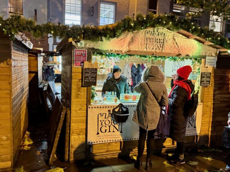 Two women stand in front of a brightly lit York Gin stall after dark at a Christmas market