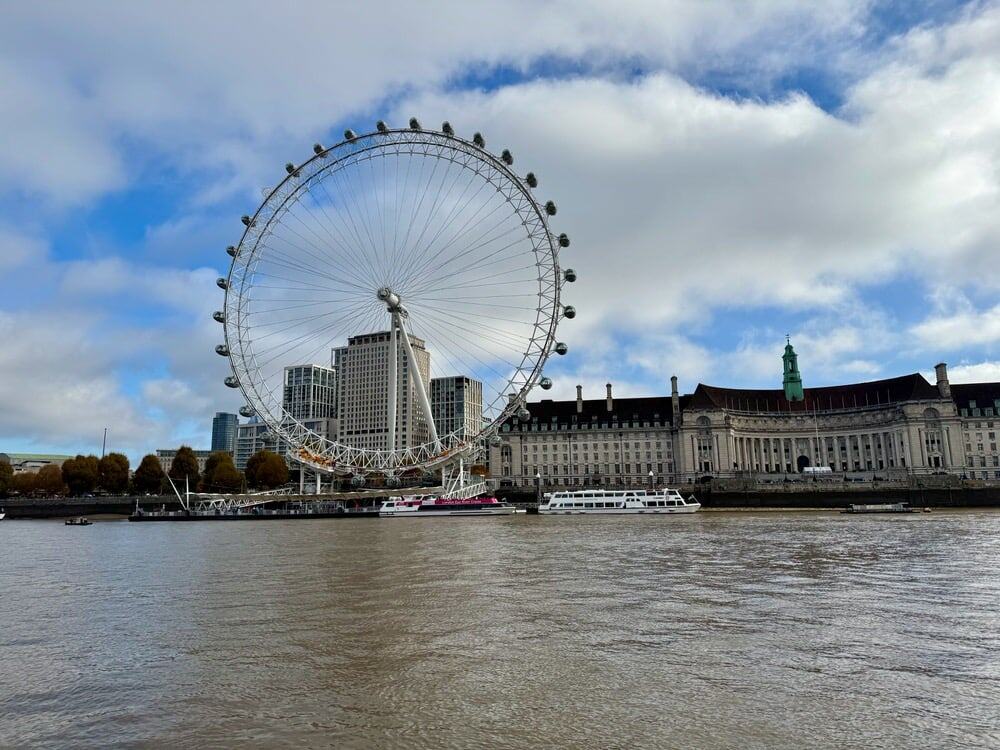 Large ferris wheel on the banks of the Thames River in central London next to a government building and river boats